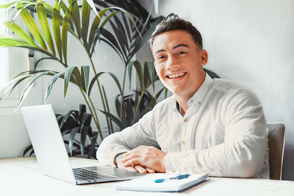 Dussmann trainee sitting at a desk in the office in front of a laptop and smiling. 