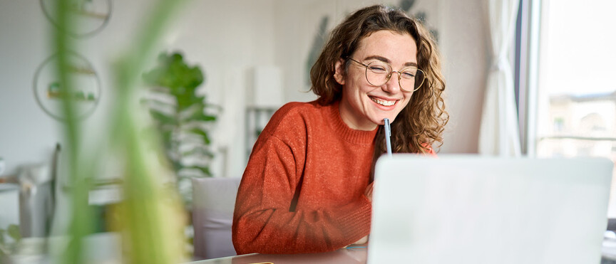 Young woman student with curly hair, wearing glasses and an orange sweater, sitting at a desk in front of a laptop and taking part in a video conference. 