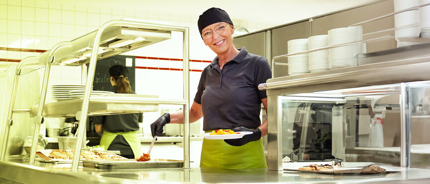 Dussmann employee from the food service and hospitality division serving food in the cafeteria and smiling.