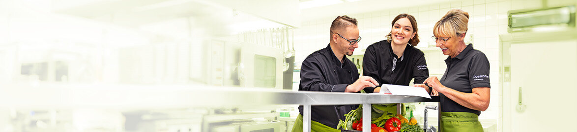 Group picture of 3 smiling Dussmann employees from the food service and hospitality division standing in an industrial kitchen and looking at a document together.