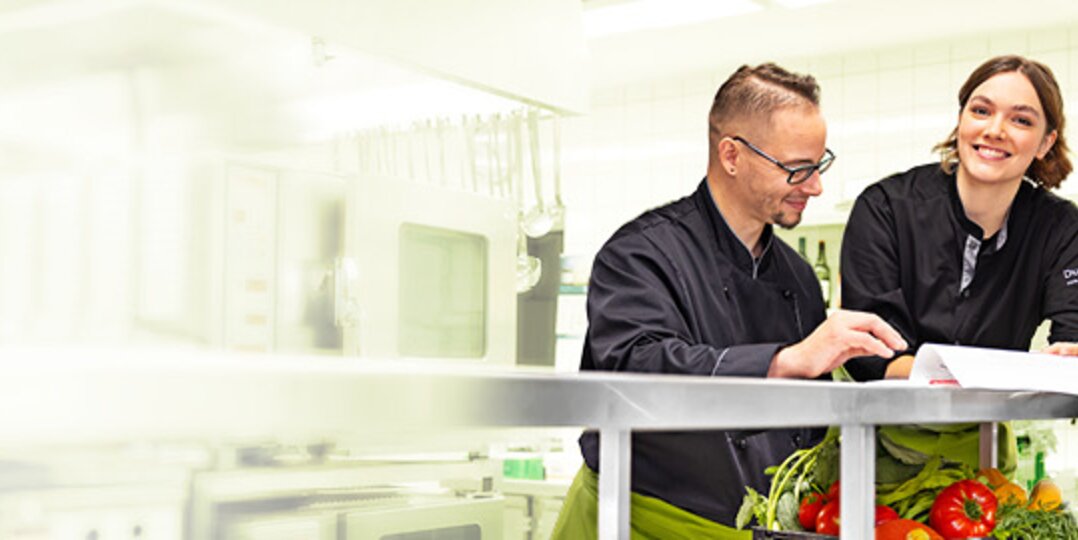 Group picture of 3 smiling Dussmann employees from the food service and hospitality division standing in an industrial kitchen and looking at a document together.