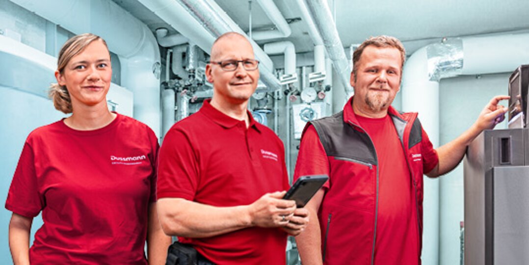 Group photo of 3 smiling Dussmann employees from the building technology and electrical engineering division in front of blue background. 