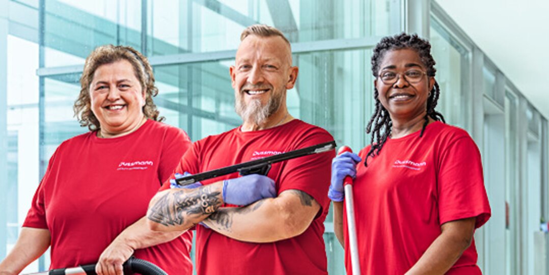 Group photo of 3 smiling Dussmann employees from the cleaning services division against a blue background. 