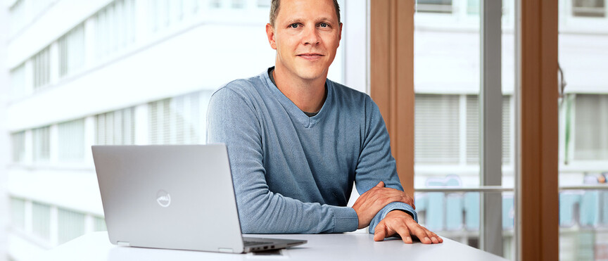 Dussmann employee from the commercial services division, sitting in an office in front of his laptop and smiling. 