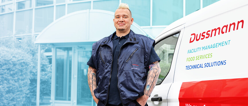 Dussmann employee from the security service division in front of his company car parked in front of a large building. 