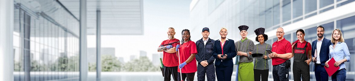 Group picture of smiling Dussmann employees from different work areas in front of a neutral gray background. 