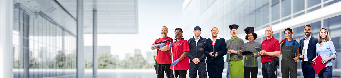 Group picture of smiling Dussmann employees from different work areas in front of a neutral gray background. 