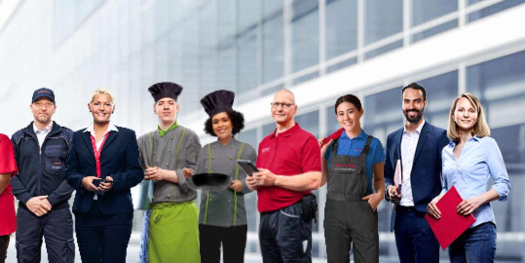 Group picture of smiling Dussmann employees from different work areas in front of a neutral gray background. 