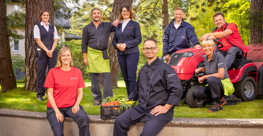 Group picture of smiling Dussmann employees from different work areas standing in a kind of large front yard with lawn and trees, wearing their work clothes. 
