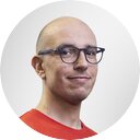 Portrait of a smiling man with a bald head, glasses and a red t-shirt in front of a grey background