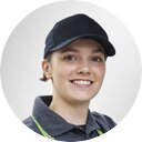 Portrait of a smiling woman with medium-length hair, dark cap, polo shirt and green apron in front of a gray background